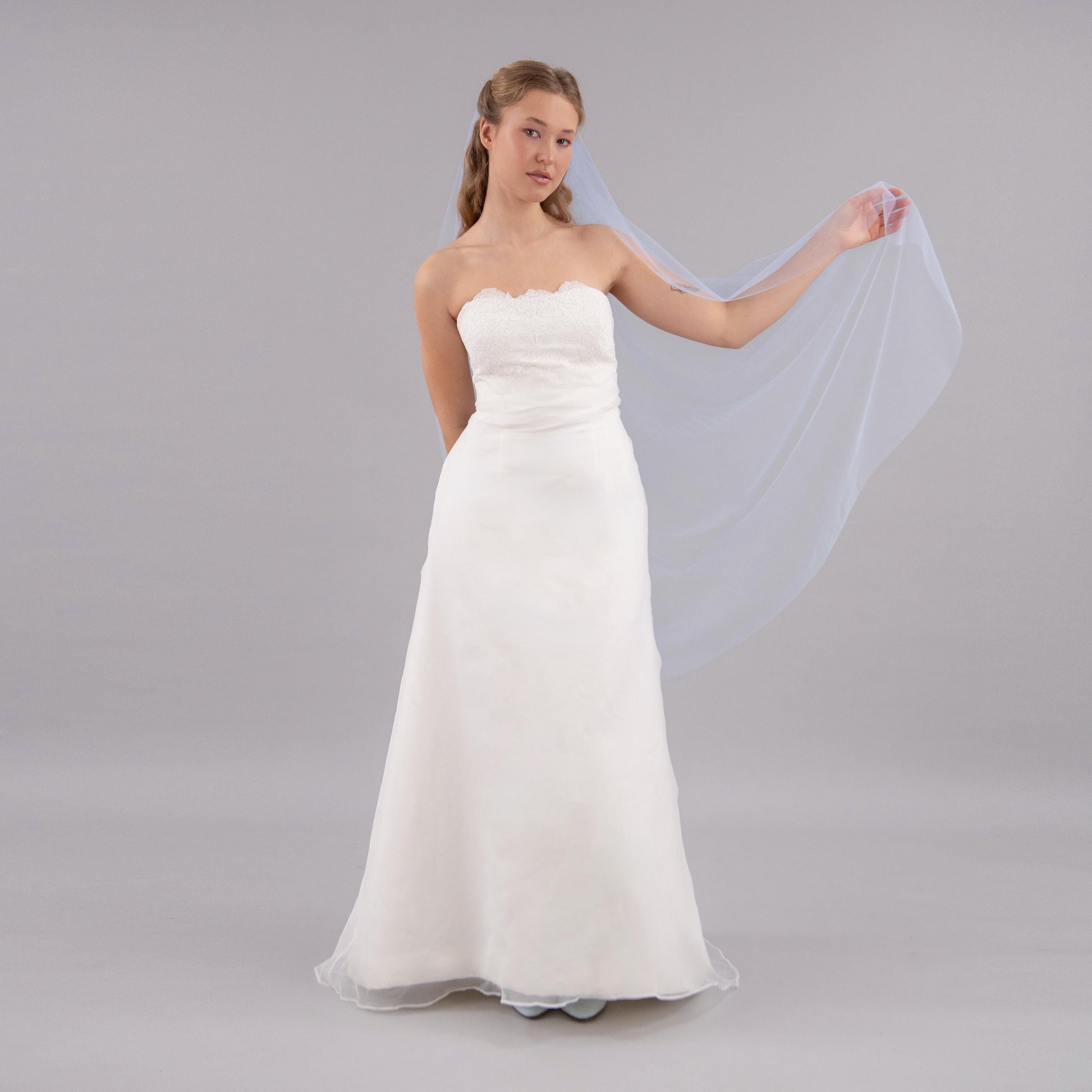 Woman wearing a white strapless wedding dress holding a pastel blue veil against a gray background