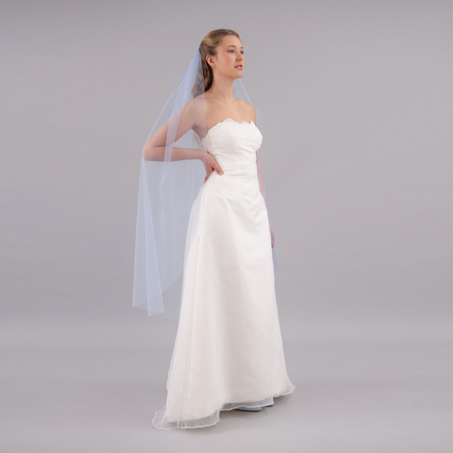 Woman wearing a strapless white wedding dress with a sheer blue veil on a gray background
