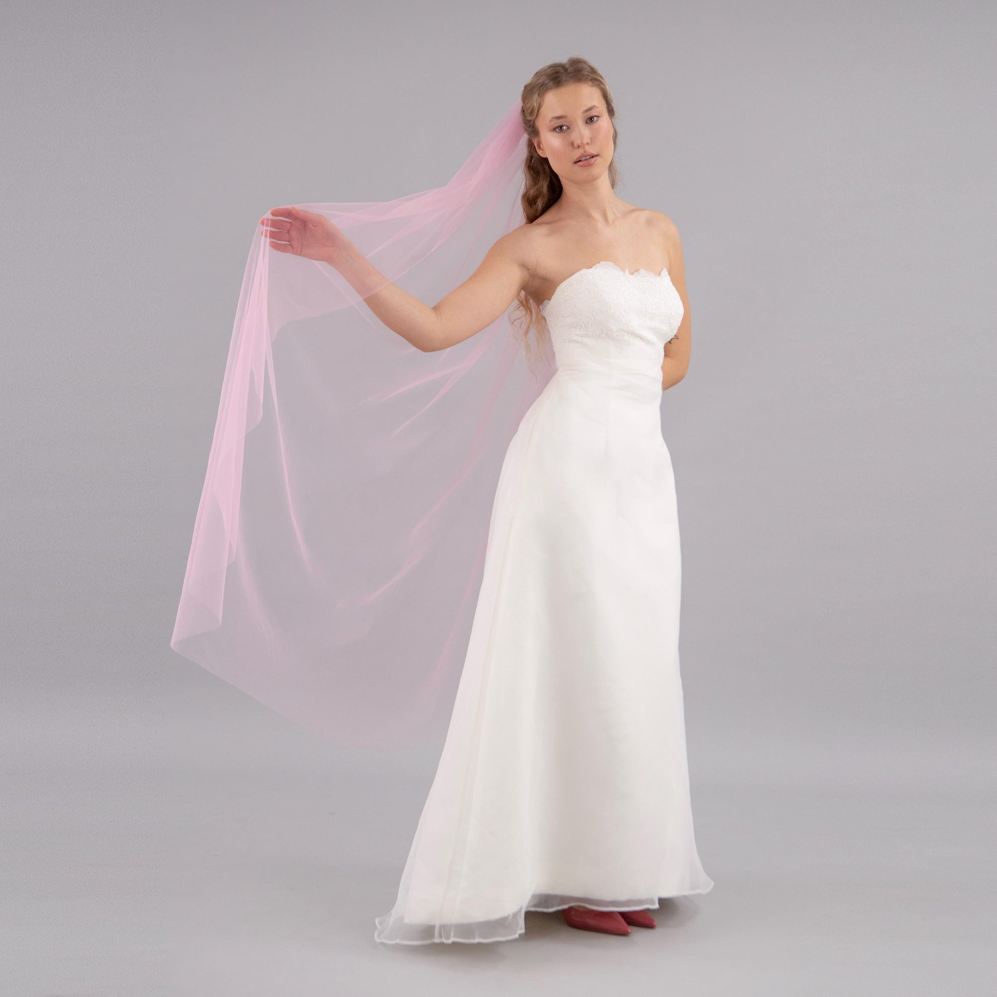 Woman in a white strapless gown holding a pink veil against a gray background